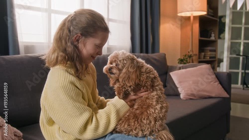 Gentle maltipoo dog licking nose of smiling gen alpha girl and looking at camera while kid sitting on couch holding pet on her lap