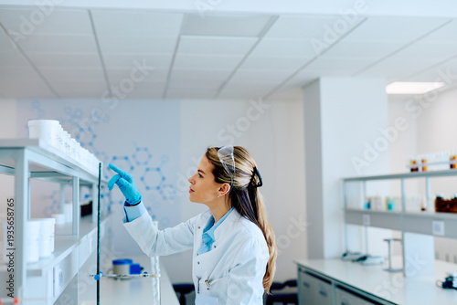 Wallpaper Mural Young female researcher wearing safety goggles and gloves, selecting chemical samples from shelves during scientific work in a lab Torontodigital.ca