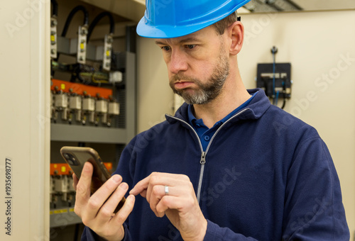 Scared, surprised man, worker in helmet after seeing instructions on mobile phone. Maintenance, circuit breaker in electrical substation