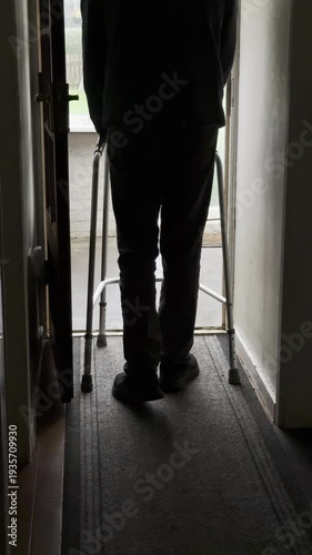 Vertical video social media - A senior man in silhouette holding a lightweight walking frame as he moves along an indoor passage or corridor, then carefully lifts the frame to go a step down.