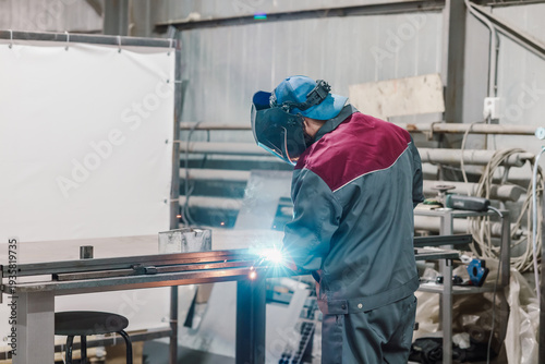 Professional welder in protective uniform and mask welding metal structures in industrial workshop.