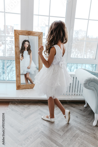Obraz na plátně A little girl in a white dress looks at her reflection in the mirror, childish dreaminess and self-perception