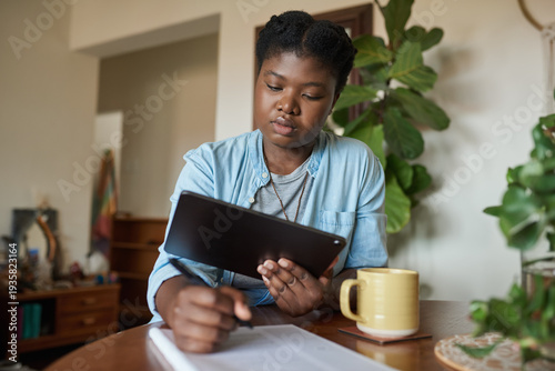 Young African woman writing notes and working on a tablet at home
