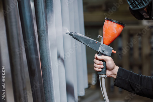 Professional worker in protective mask using powder coating spray gun on metal pipes in factory.