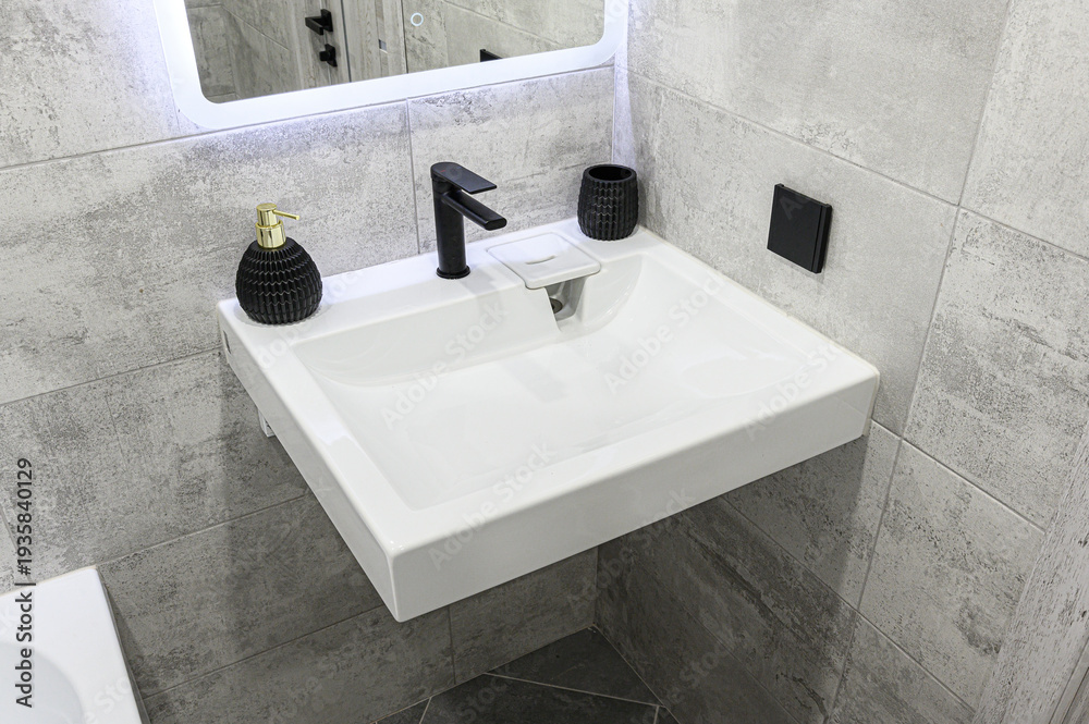 Fototapeta premium Modern white wall-mounted sink with black faucet; grey textured tiles, illuminated mirror, black accessories (soap dispenser, cup), sleek minimalist bathroom design