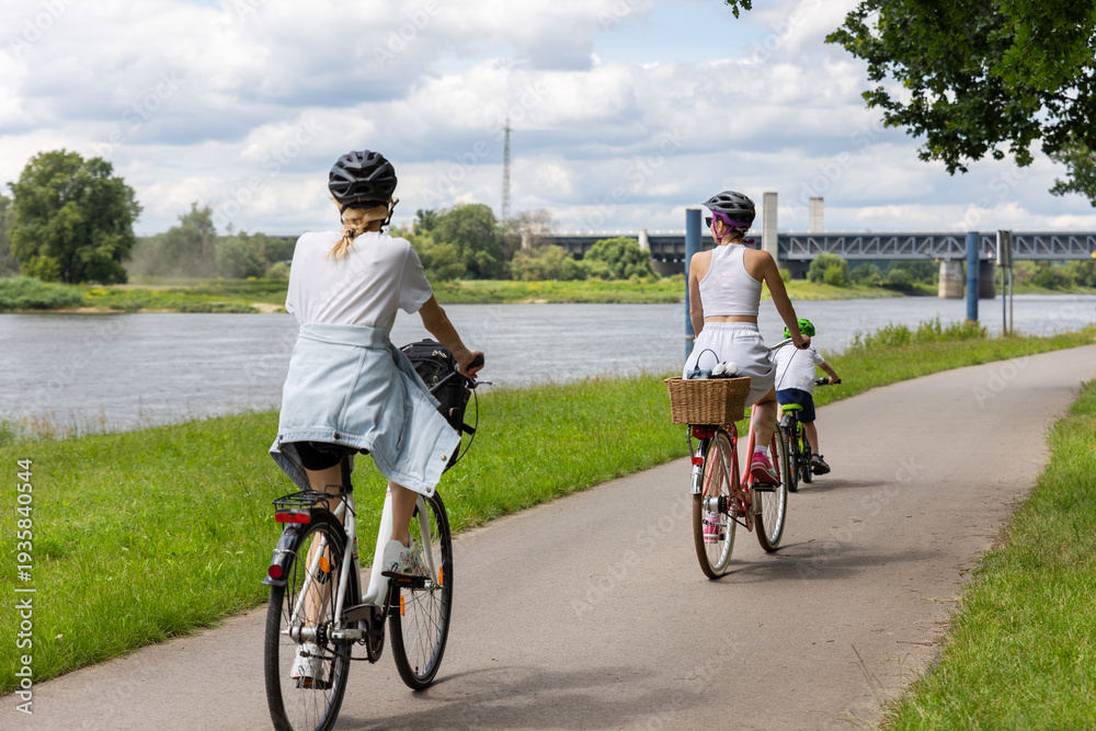 Fototapeta premium Family cycling along riverside bike path near Elbe in Magdeburg under dramatic clouds. Mother child riding bicycles helmets through green landscape symbol healthy lifestyle freedom summer recreation