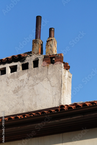 Low angle shot of a weathered concrete chimney with metal pipes on a traditional tiled roof against a clear blue sky