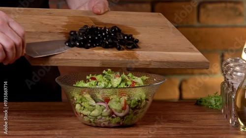A male chef slices black olives for a salad on a cutting board and mixes the salad in a bowl.