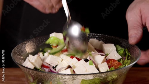 The cook tosses the salad with white feta cheese, tomatoes, olives and lettuce leaves in a bowl with a spoon.