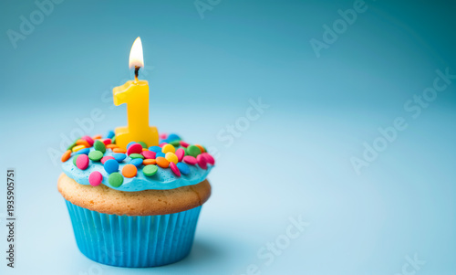 Cupcake with blue icing, colorful candy sprinkles, and a yellow number one candle, presented on a blue background, first birthday treat, cheerful party, milestone celebration.
