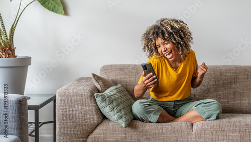 Excited happy young black woman holding smart phone device sitting on sofa at home - Happy satisfied female looking at mobile smartphone screen gesturing yes with clenched fist - Technology concept