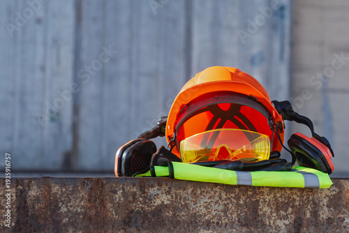 Orange hard hat with protective goggles, earmuffs and high visibility vest against industrial background. Personal protective equipment for construction workers and industrial safety.