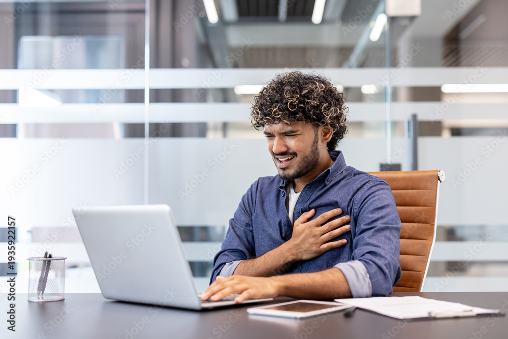 Fototapeta premium A young Indian man, twisted in pain, sits in an office at a desk with a laptop and holds his hand to his chest