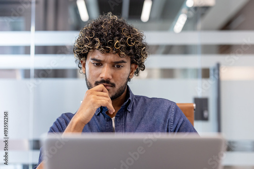 Thoughtful and focused young Indian man working in the office on a laptop