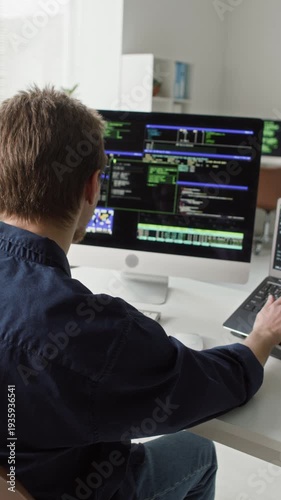 Vertical shot of focused young adult male IT developer in glasses typing on laptop and desktop computer at programming desk writing software code in modern office