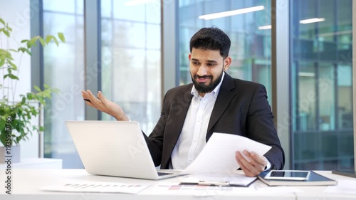 Confused frustrated businessman having difficulty with paperwork sitting at workplace in business office. Puzzled male worker in suit looks at documents and laptop screen and can't understand problem.