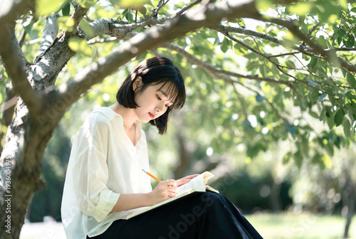 Young asian woman sitting outdoors on a bench and writes in notebook in dappled sunlight. Peaceful lifestyle style suitable for education, mental health, and leisure branding.