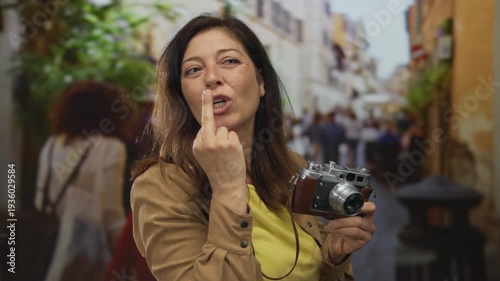 Hispanic middle age woman holding vintage camera shows middle finger with a grin on busy street; defiance.