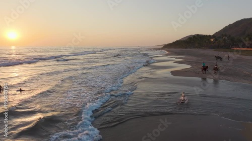 Aerial view of dog running along ocean shoreline waves at sunset as horseback riders pass along the beach