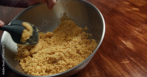 Mixing crushed cookies with melted butter in metal bowl to form cheesecake crust. Close up of homemade dessert base preparation on wooden table in kitchen.