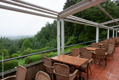 Empty restaurant terrace with wicker chairs and tables overlooking a lush green forest valley on a rainy day in Vaals