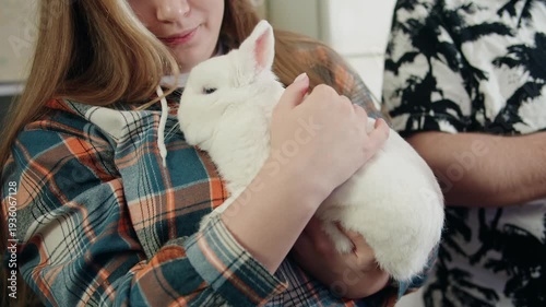 Young caucasian woman in plaid hoodie cradling and gently petting white relaxing rabbit with soft fur in cozy kitchen. Closeup of nurturing caretaking and grooming of pets