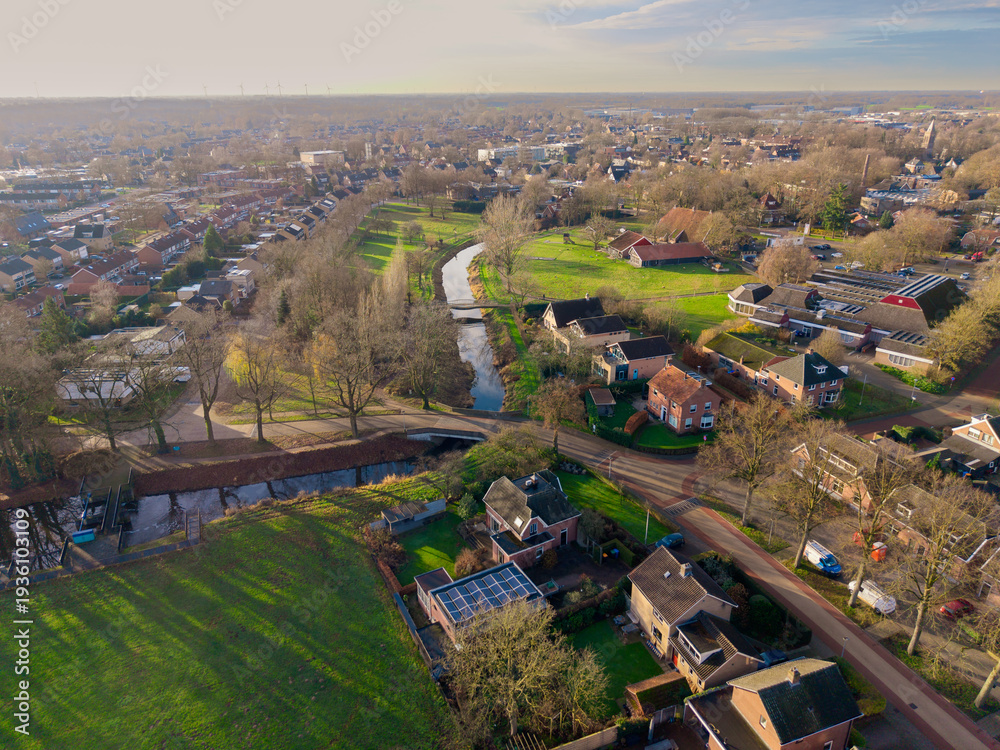 Fototapeta premium Aerial view of a tranquil European town with a winding canal, green parks, and brick houses under a soft morning sun. A peaceful suburban landscape with traditional architecture and nature.