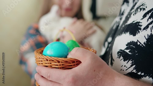 Caucasian young woman hugging and cradling white sleeping rabbit while man holding basket of painted easter eggs on foreground in kitchen. Spring easter tradition and warm family exchange