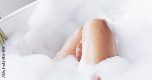 Lower legs are rising from foam in bathtub as subtle leg movement is parting bubbles, repositioning