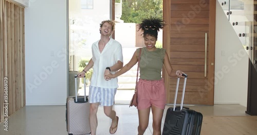 Diverse couple entering glass door opening, pulling suitcases, hugging and returning home