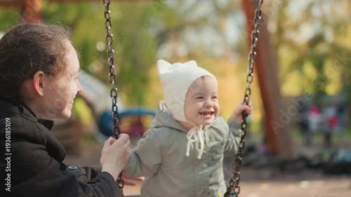 Closeup of joyful toddler laughing on swing with mother nearby, autumn park backdrop, caucasian parent celebrating childs milestone, knit hood and coat details, warm golden light, authentic family