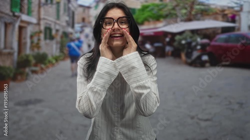 Young hispanic woman wearing glasses cups hands to mouth on cobblestone street lined with buildings; excitement.