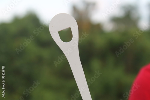 Close-up of a white plastic object shaped like a spoon or tag with a hole, isolated against a soft blurred green nature background. Minimalist photography focusing on shape, texture, and bright natura