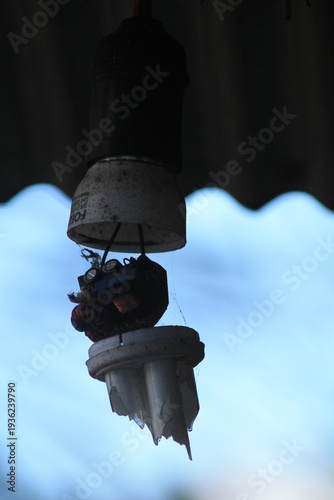 Broken and dusty energy-saving CFL light bulb hanging from a dark ceiling. Concept of power outage, electricity failure, waste, and neglected rural infrastructure with a blue sky background.
