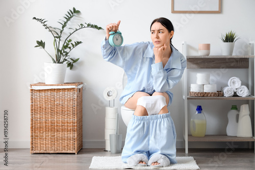 Displeased young woman with alarm clock sitting on toilet bowl in restroom