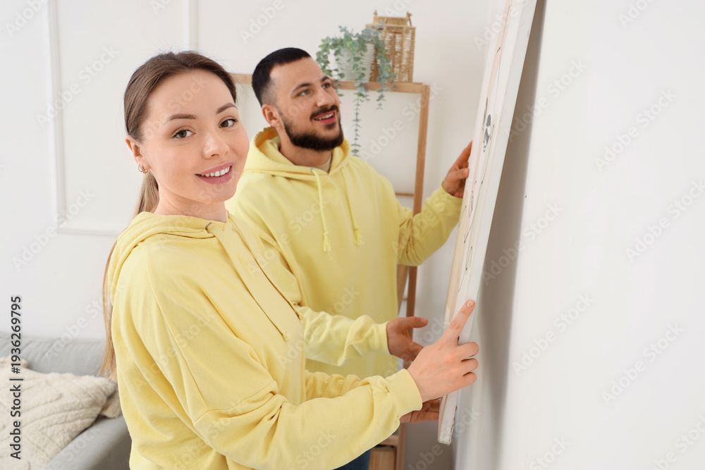 Fototapeta premium Young couple hanging painting on light wall at home