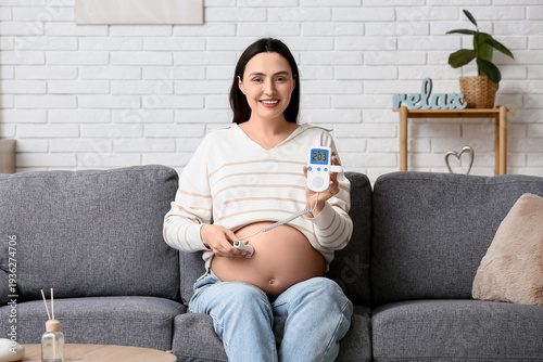 Young pregnant woman with fetal doppler listening to baby's heartbeat on sofa...