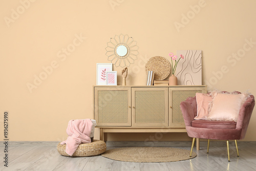 Interior of stylish room with pink armchair, wicker ottoman and home decor on commode near beige wall