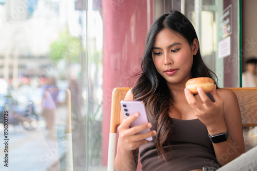 Young Woman Looking at Smartphone While Holding Donut in Cafe