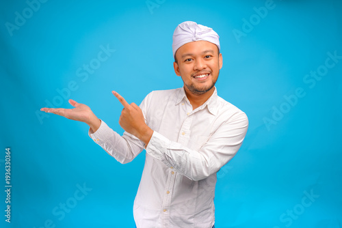 Smiling Balinese man wearing traditional Indonesian udeng pointing to the copy space on beside him.