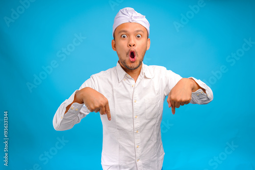 Shocked Balinese man wearing Indonesian traditional udeng pointing up to the copy space downwards.