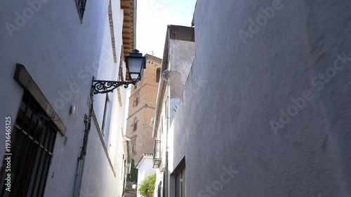Wallpaper Mural POV walking shot through a narrow alley in the Albaicin district, revealing the Mudejar tower of San Bartolome Church. Bright daylight, white facades, and traditional Spanish atmosphere. Torontodigital.ca
