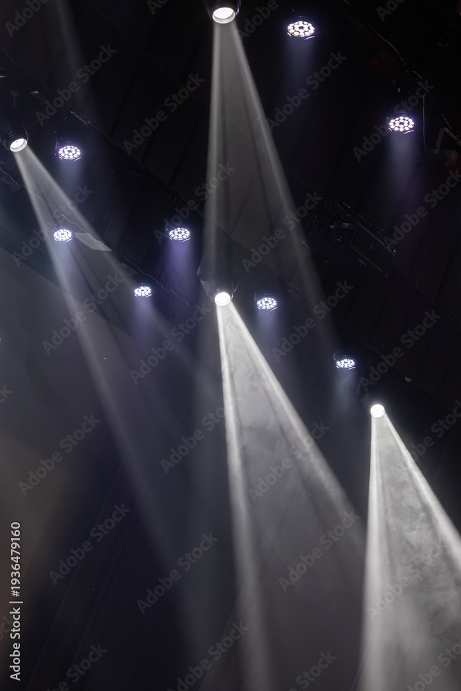 Naklejka premium Bright stage spotlights shine downward through swirling theatrical smoke in a dark performance venue, creating dramatic beams of light with strong contrast and atmospheric lighting