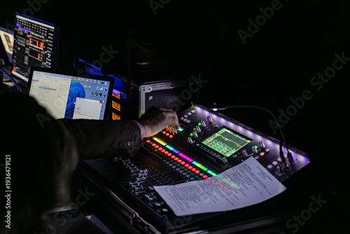 A sound engineer adjusts controls on a digital audio mixing console surrounded by illuminated buttons and screens in a dark control area during a live event or concert production