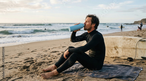 Surfer drinking from bottle sitting by surfboard on beach