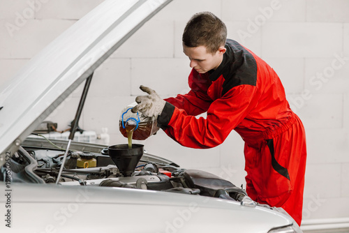 Professional car mechanic in red uniform pouring new engine oil from a plastic bottle into a vehicle engine. Auto technician performing maintenance service in a modern workshop garage.