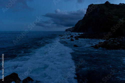 A rocky shoreline with a dark sky and ocean waves. there is a lighthouse in the distance.