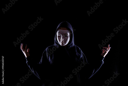 Foto Person wearing a white expressionless mask and dark hoodie standing against a black background with raised hands in a dramatic gesture