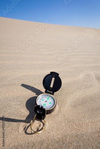 A navigation compass rests on a sandy dune beneath a vibrant blue sky, representing themes of travel, exploration, navigation, and adventure in a desert or coastal environment.