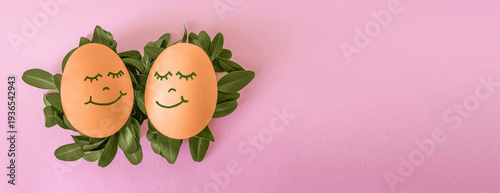 Easter spring banner (card): two natural brown eggs with painted smiling faces, lying on green fresh leaves on a delicate pink background